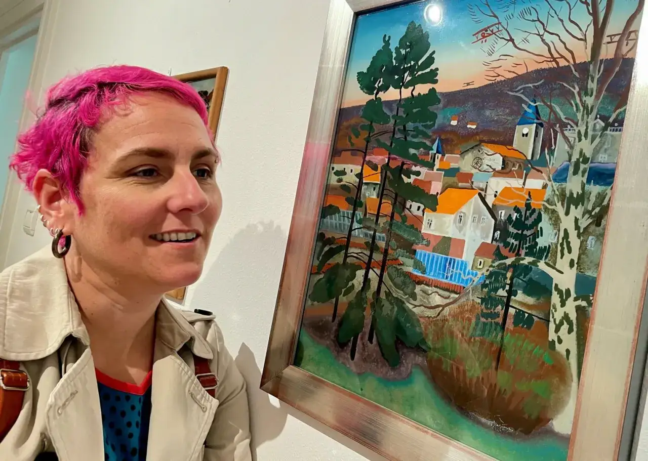 Une femme aux cheveux roses admire un tableau d'&Eacute;tienne Cournault repr&eacute;sentant un village pittoresque sous un ciel color&eacute;.