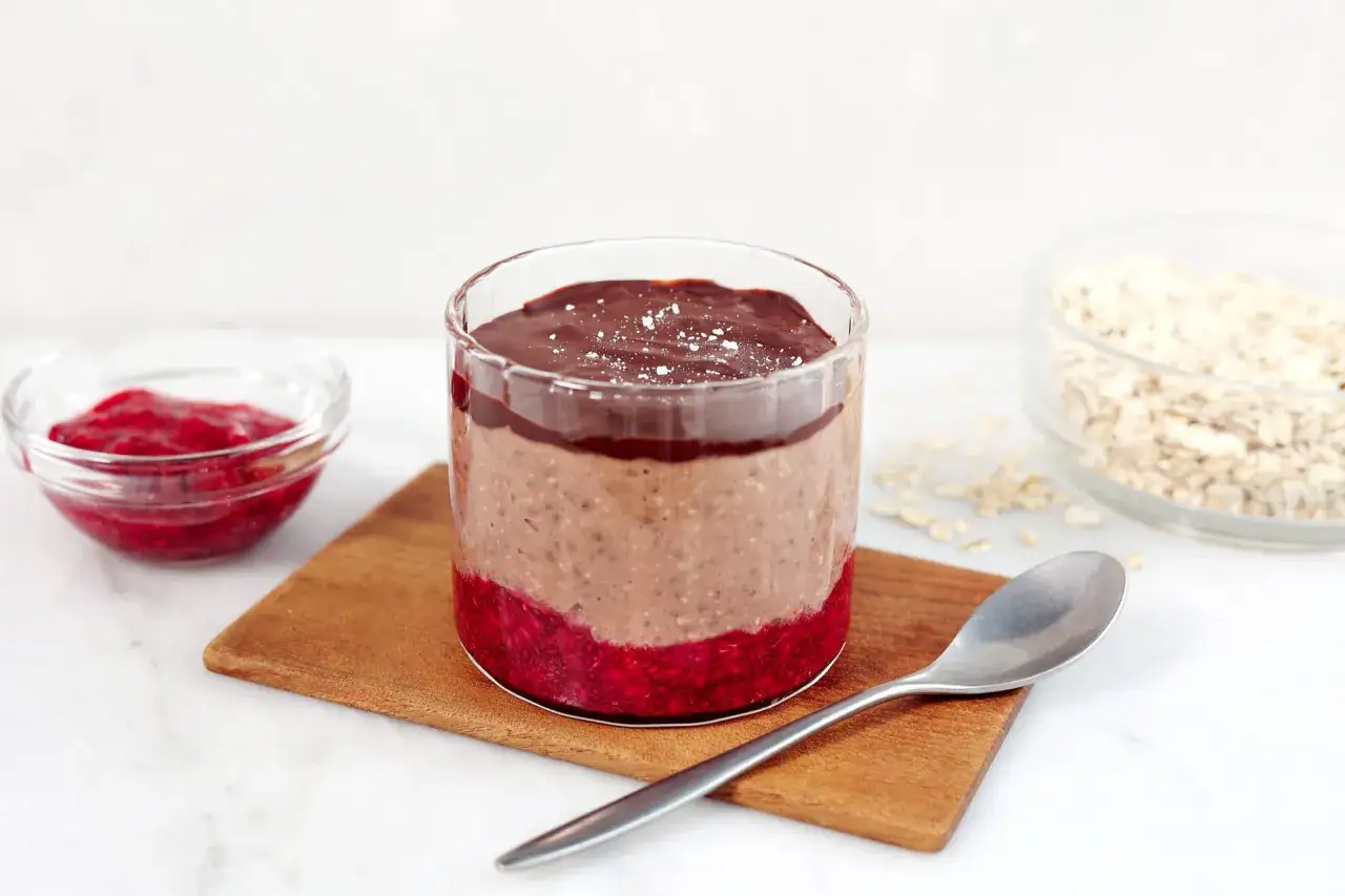 En glasburk med overnight oats med chokladsmak, hallonsylt och en sked. Bredvid finns en skål med hallon och en skål med havregryn.