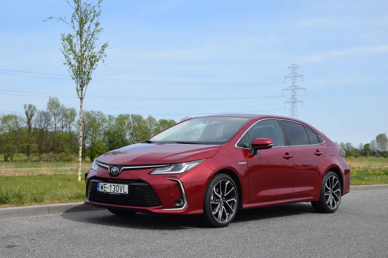 Czerwona Toyota Corolla 1.8 Hybrid zaparkowana przy drodze, w tle drzewa i słup energetyczny.