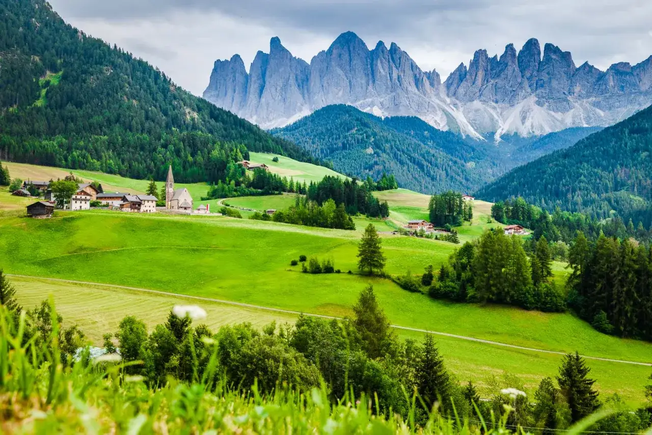 Grüne Hügel und traditionelle Häuser im **Val di Funes**, mit den schroffen Dolomiten im Hintergrund.