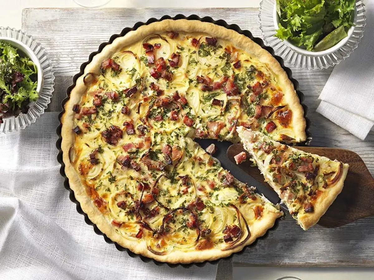 Herzhaftes Quiche Rezept mit Speck und Zwiebeln, frisch aus dem Ofen. Ein St&uuml;ck wird gerade serviert.
