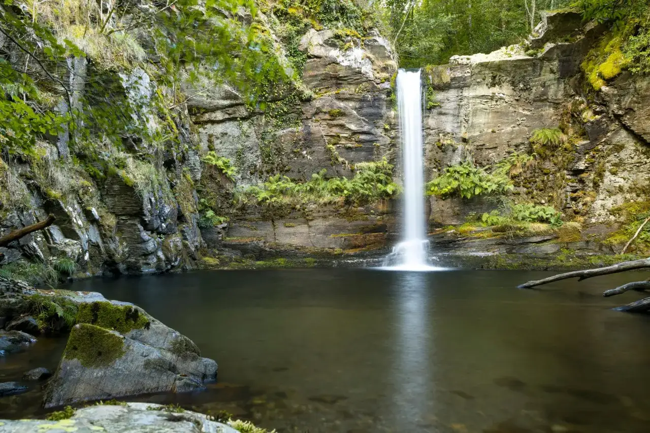 La **fervenza do Pombar** cae en cascada sobre rocas cubiertas de musgo, creando un remanso de paz en medio de la naturaleza exuberante.
