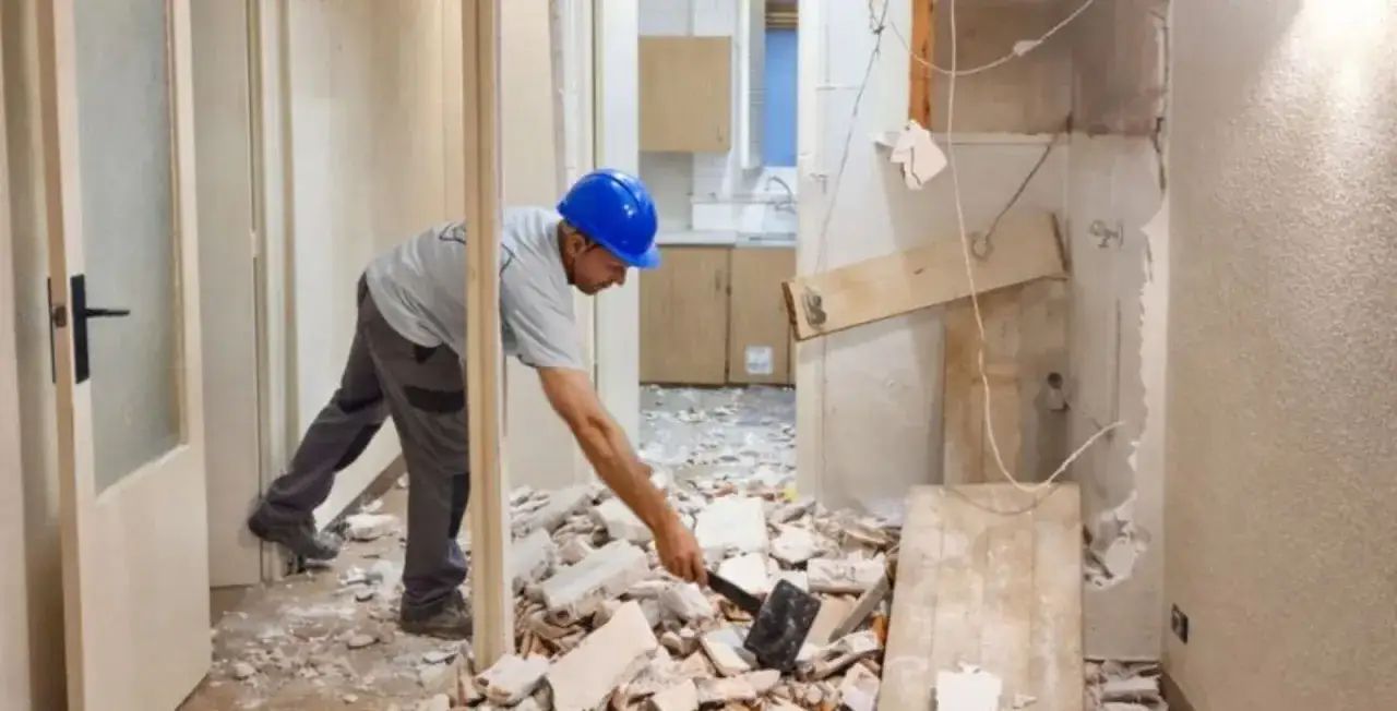 Obrero con casco azul trabajando en la demolici&oacute;n de un tabique, rodeado de escombros.