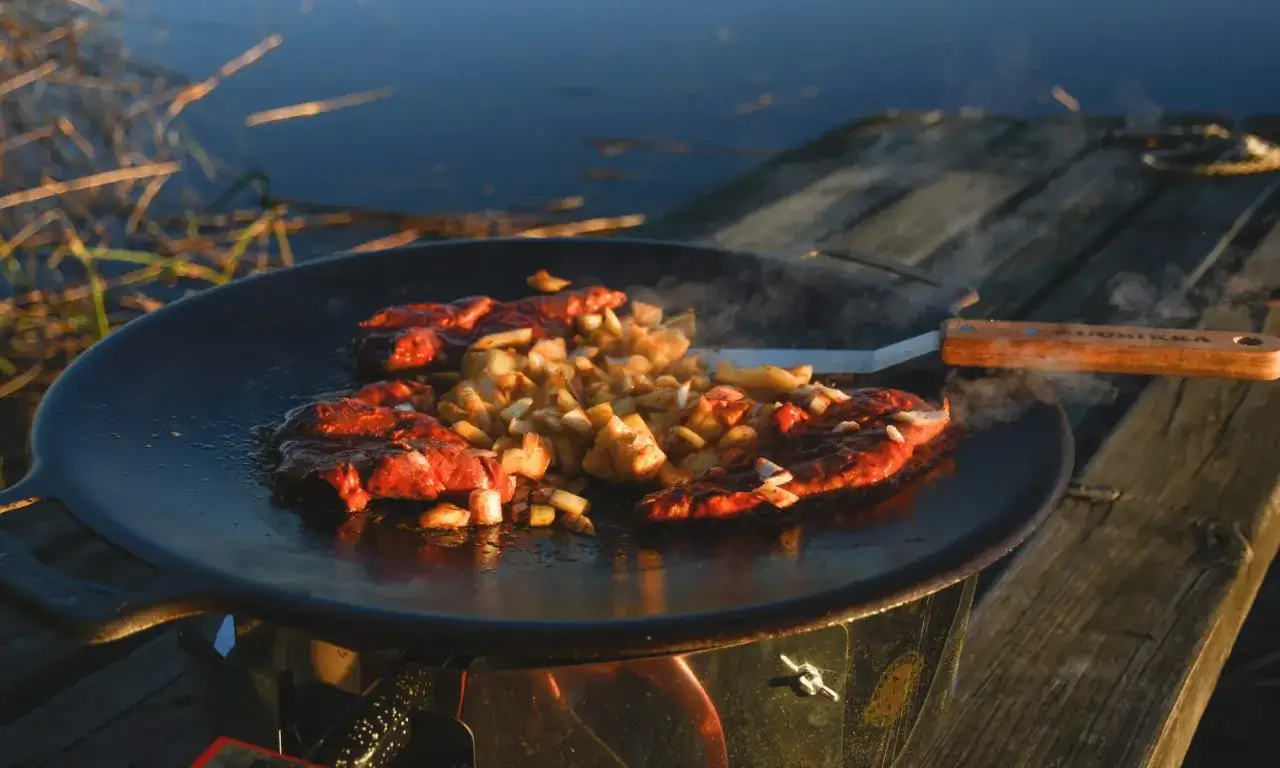 K&ouml;tt och potatis steks p&aring; en rostig muurikka vid vattnet.