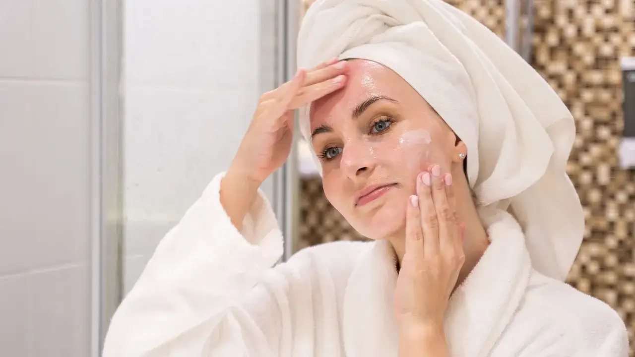 Mujer con toalla en la cabeza y bata, aplicándose crema. Descubre las mejores cremas antiedad 60 años ocu para una piel radiante.