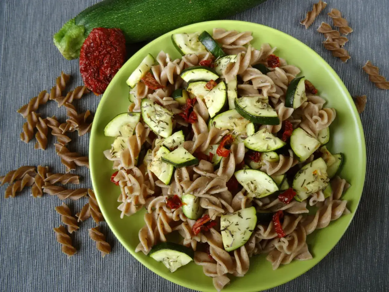 Pyszny makaron z cukinią i pomidorami, posypany ziołami. Obok leżą surowe składniki i makaron fusilli.
