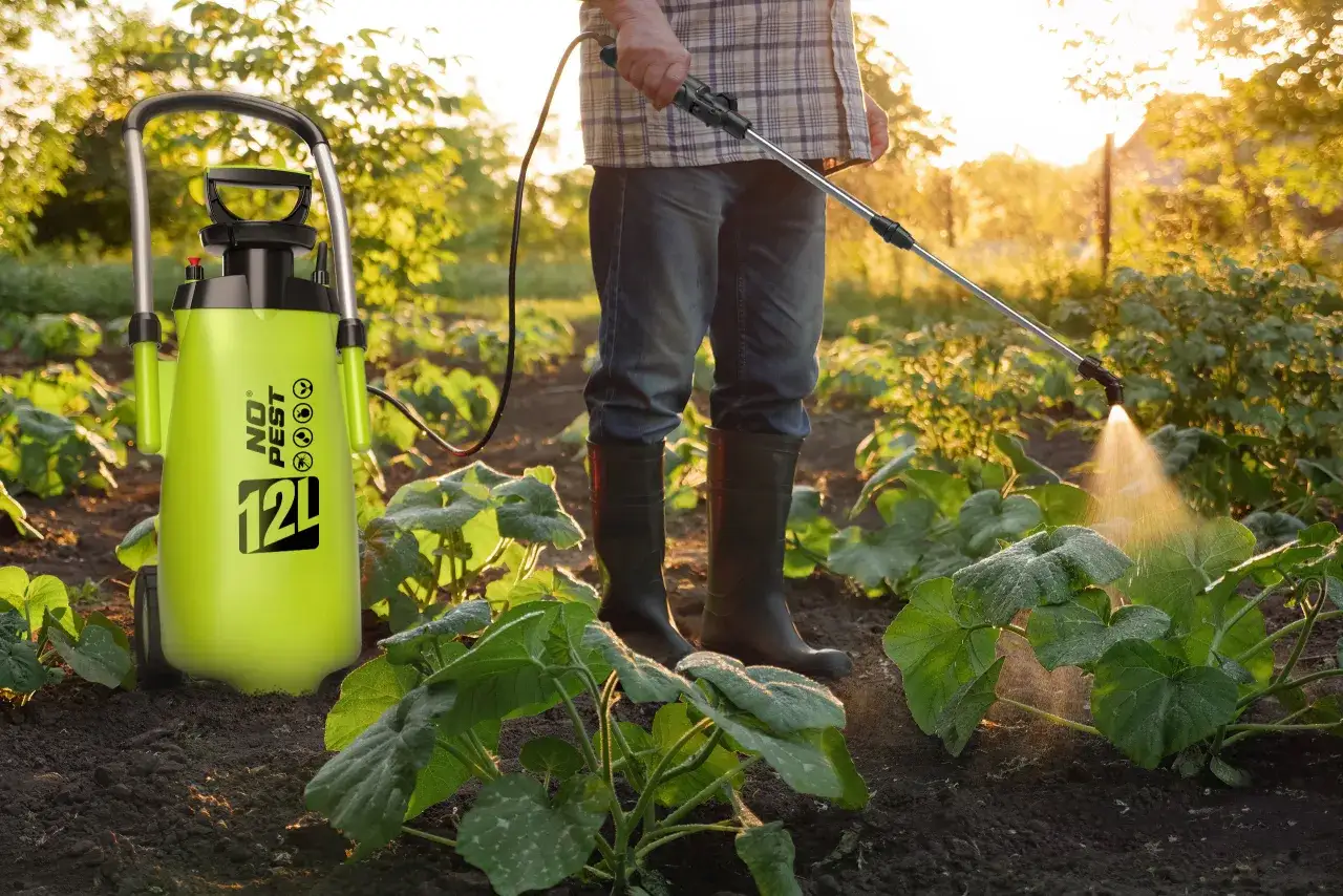 Zielony opryskiwacz do trawnika o pojemności 12L stoi obok grządek, a mężczyzna w gumowcach opryskuje rośliny.