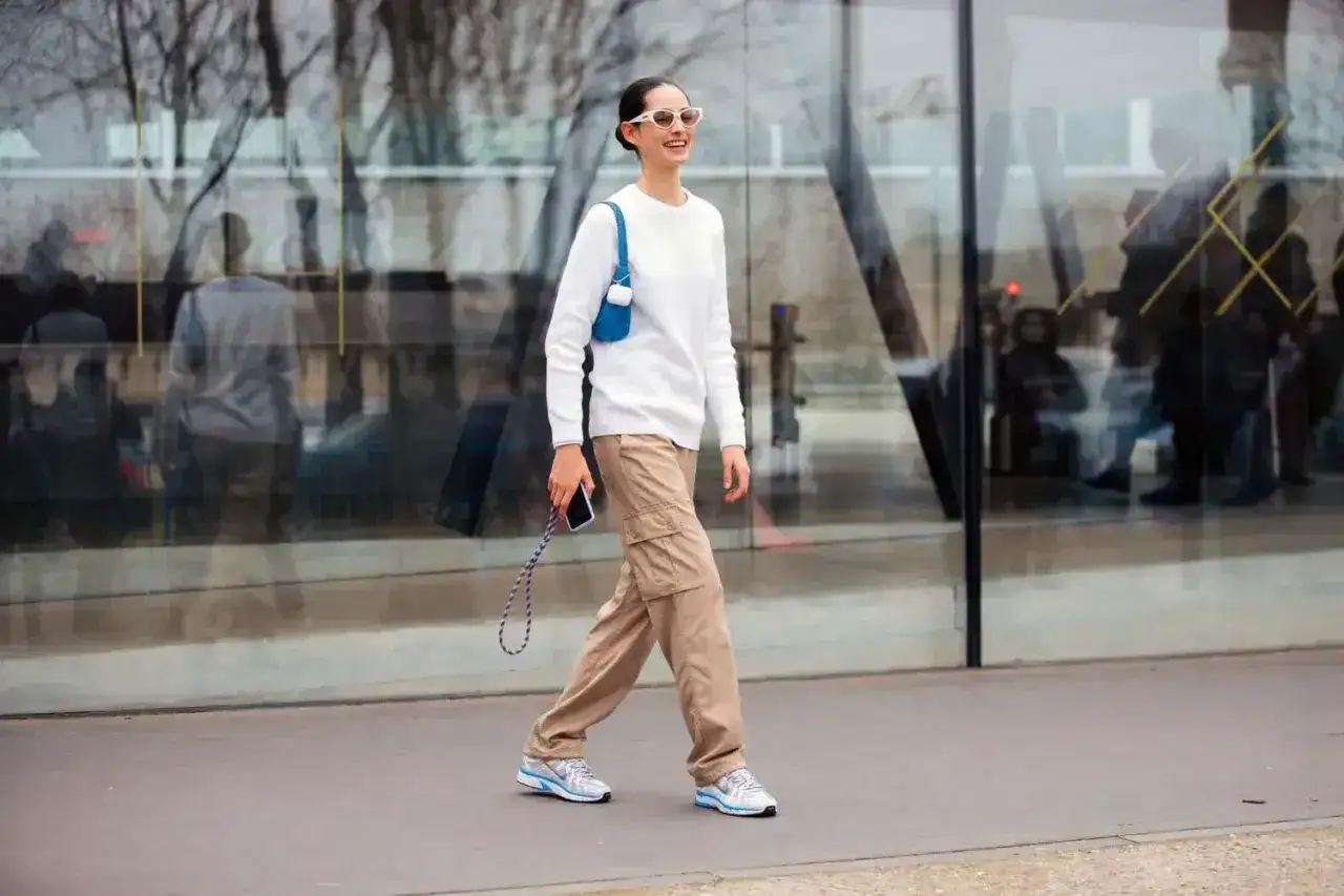 Kobieta w białym swetrze i spodniach cargo w kolorze khaki idzie ulicą, trzymając telefon.