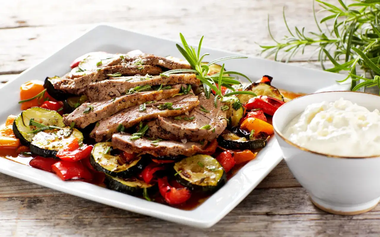 Skivad lammstek i ugn serverad med ugnsrostade gr&ouml;nsaker som zucchini, paprika och tomater. En klick vit s&aring;s i en sk&aring;l.