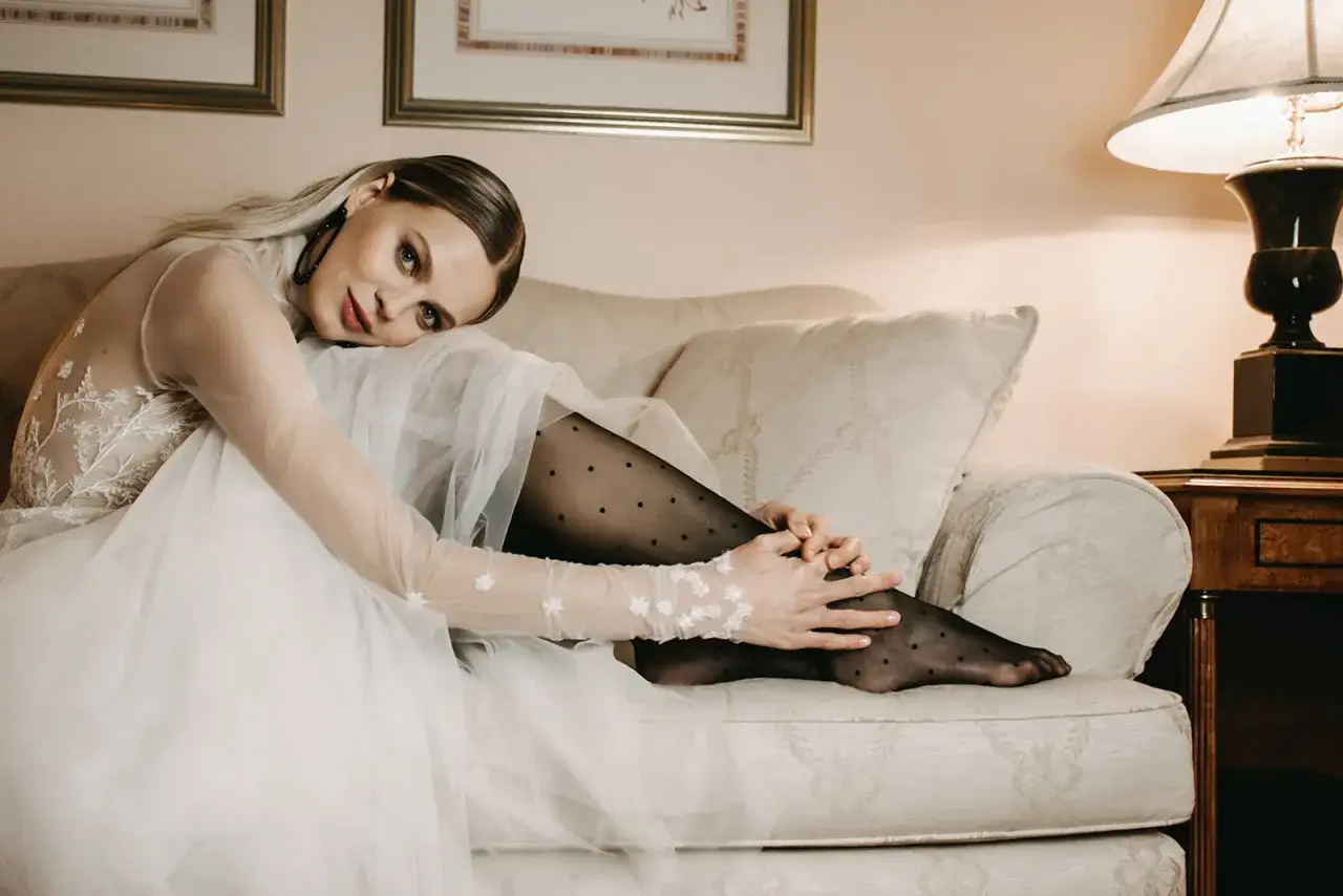 Braut mit zarter Spitze und gepunkteter Strumpfhose unter dem Kleid, die auf einem Sofa sitzt. Eine elegante Wahl, welche Strumpfhose unter Brautkleid passt.