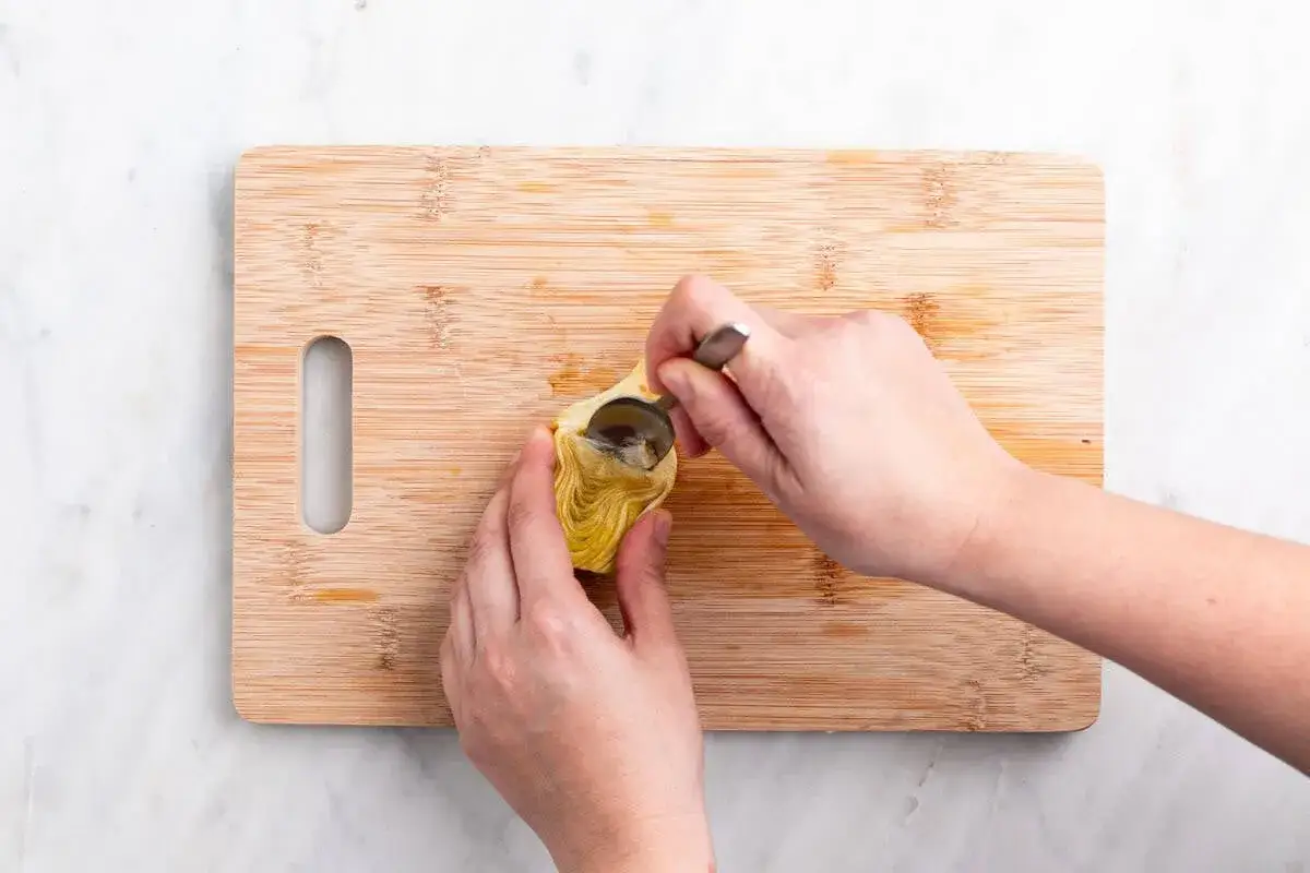 Manos preparan alcachofas a baja temperatura, extrayendo el coraz&oacute;n con una cuchara sobre una tabla de madera.