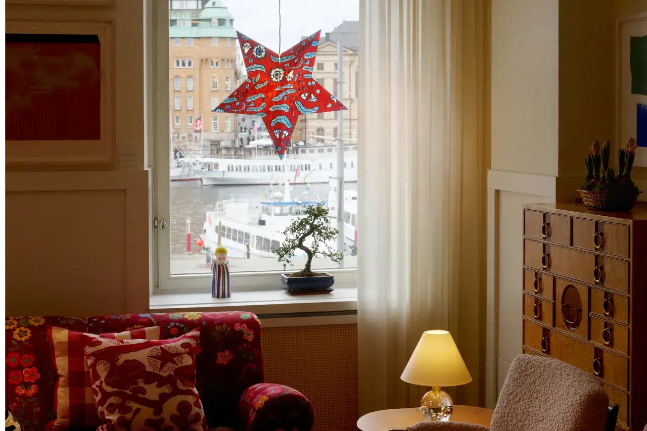 En r&ouml;d adventsstj&auml;rna med blommor h&auml;nger i f&ouml;nstret. Utanf&ouml;r syns b&aring;tar och byggnader. En bonsai och en figur st&aring;r p&aring; f&ouml;nsterbr&auml;dan.