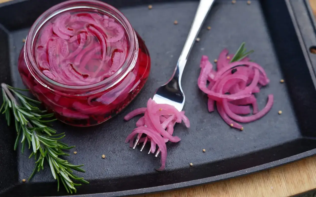Ein Glas mit leuchtend pinken, gepickelten Zwiebeln, daneben eine Gabel mit Zwiebelringen und ein Rosmarinzweig.