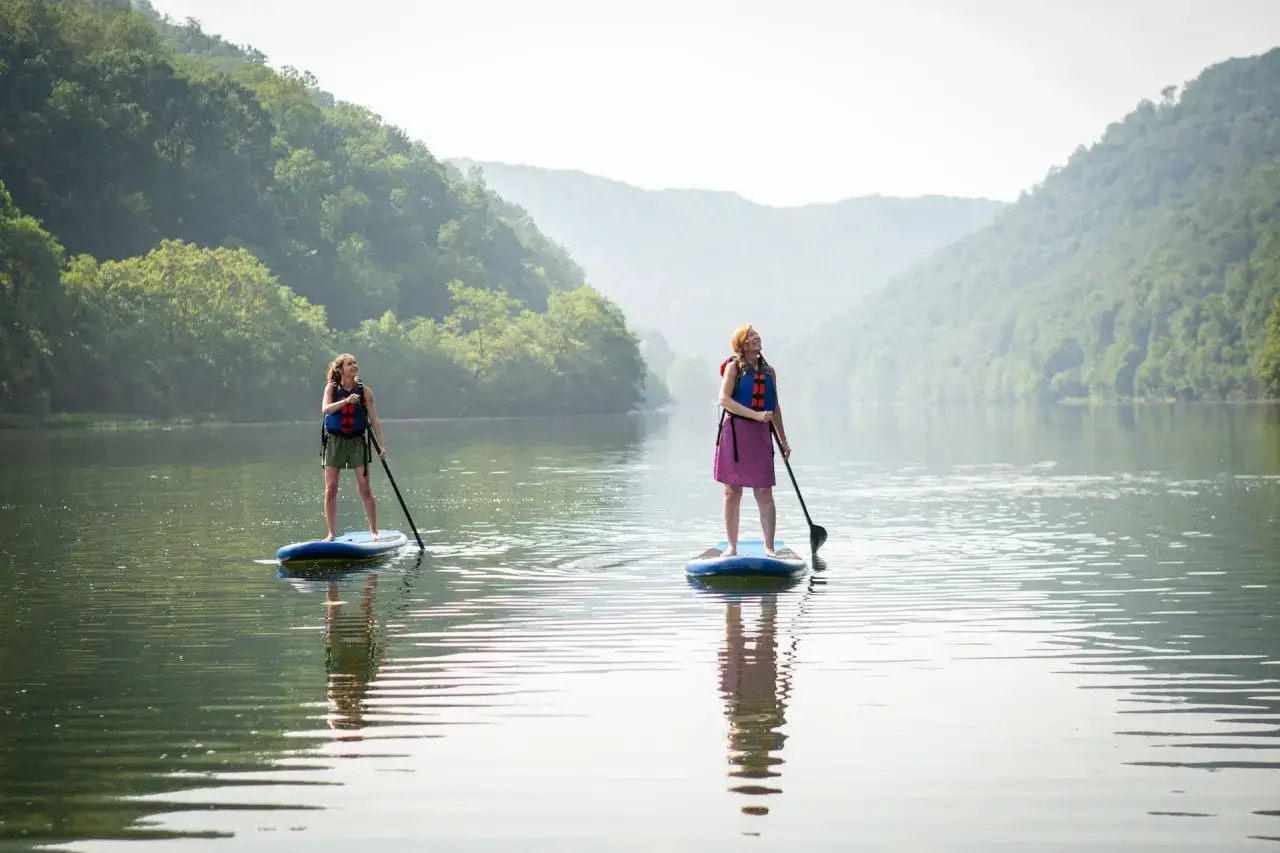 Zwei Frauen genießen eine entspannte SUP-Tour auf einem ruhigen Fluss, umgeben von sanften, bewaldeten Hügeln.