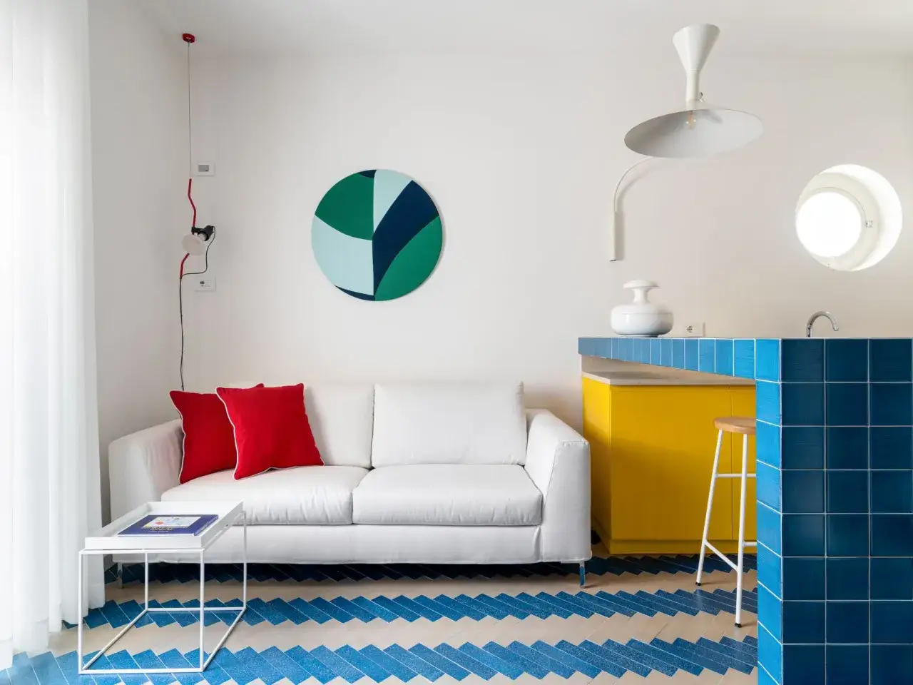 Sof&aacute; blanco, cojines rojos y suelo con patr&oacute;n de espiga azul y blanco. Unos trucos para decorar espacios peque&ntilde;os: la pared con arte circular y la barra de cocina amarilla y azul.