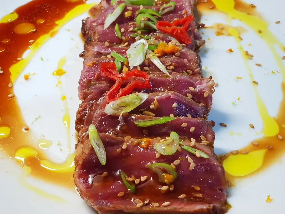 Tataki de ternera, finas lonchas de carne sellada, cubiertas de s&eacute;samo, cebolleta y chile, ba&ntilde;adas en una salsa dorada.