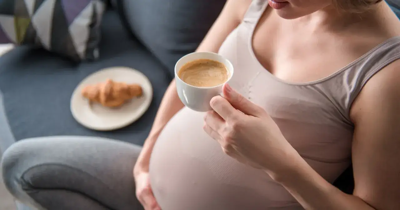 Kobieta w ciąży pije kawę rozpuszczalną, obok leży croissant.