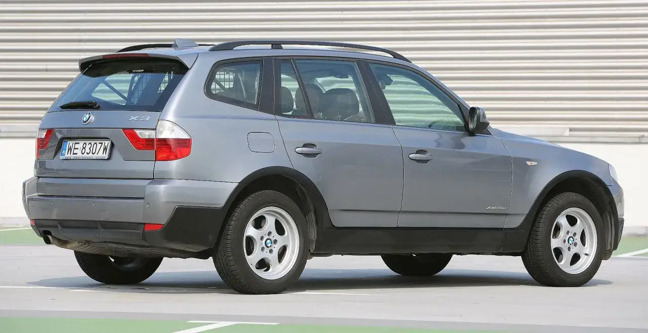 Szare BMW E83, model X3, stoi na parkingu. Widoczny tył i bok pojazdu z tablicą rejestracyjną WE 8307W.