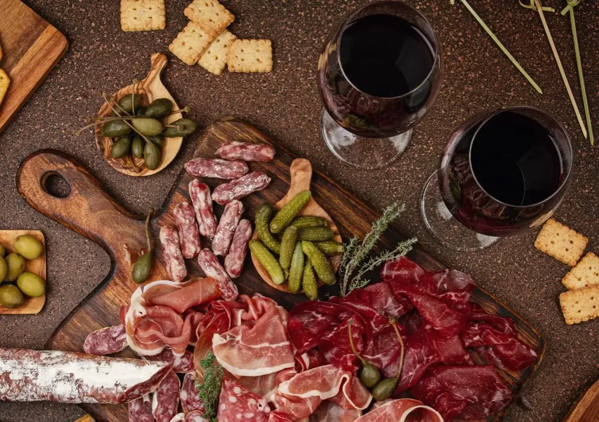 Plateau de charcuterie variée, cornichons, olives, câpres et deux verres de vin rouge. Idéal pour savoir quel vin avec charcuterie.