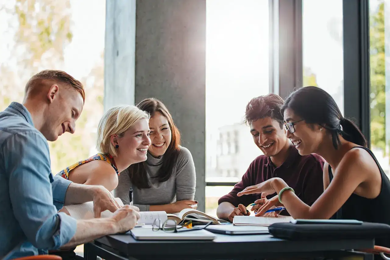 Glada studenter diskuterar under en socialpedagog utbildning distans. De sitter runt ett bord med böcker och anteckningar.