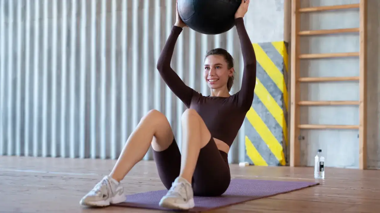 Femme souriante faisant un exercice de gainage abdos avec un ballon médicinal, sur un tapis de yoga dans une salle de sport.