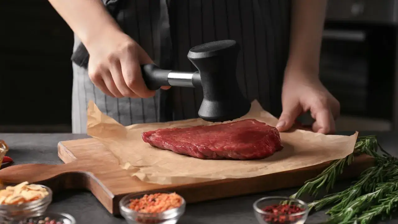 Une main utilise un attendrisseur pour aplatir un steak. C'est une étape clé pour comprendre pourquoi taper la viande rend les plats plus tendres et savoureux.
