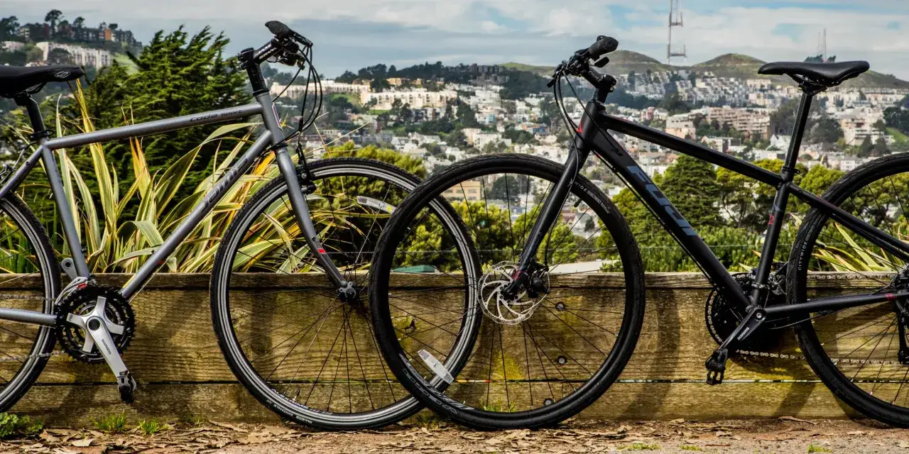 Two hybrid bikes lean against a wooden fence with a city skyline in the background. A hybrid bike is good for commuting and light trails.