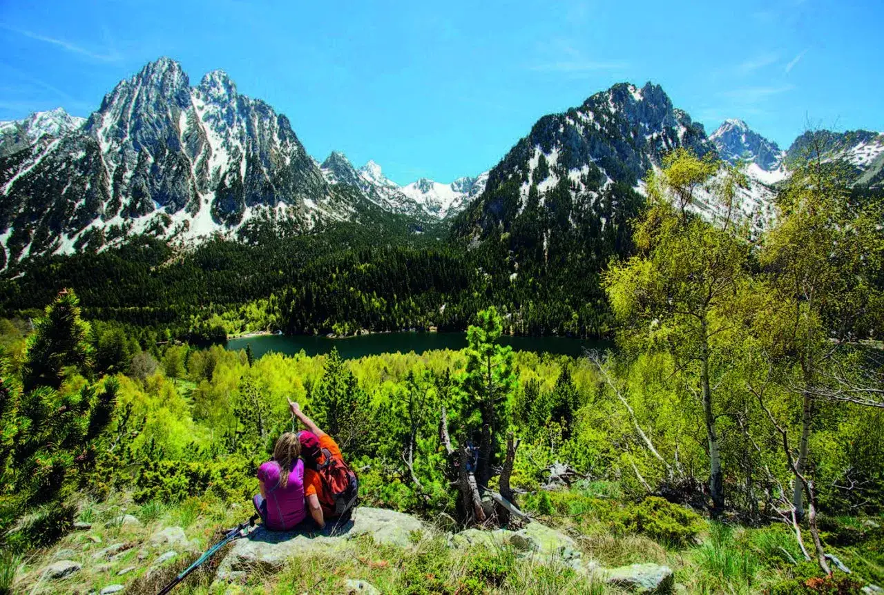 Dos excursionistas contemplan el impresionante Pirineo de Lleida, con picos nevados, un bosque frondoso y un lago sereno.