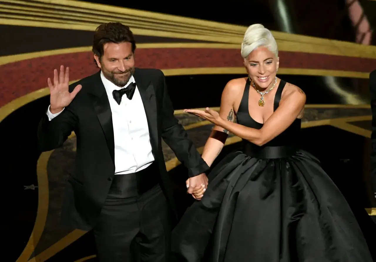 Bradley Cooper i Lady Gaga trzymają się za ręce na scenie. Bradley w czarnym smokingu, Lady Gaga w czarnej sukni.