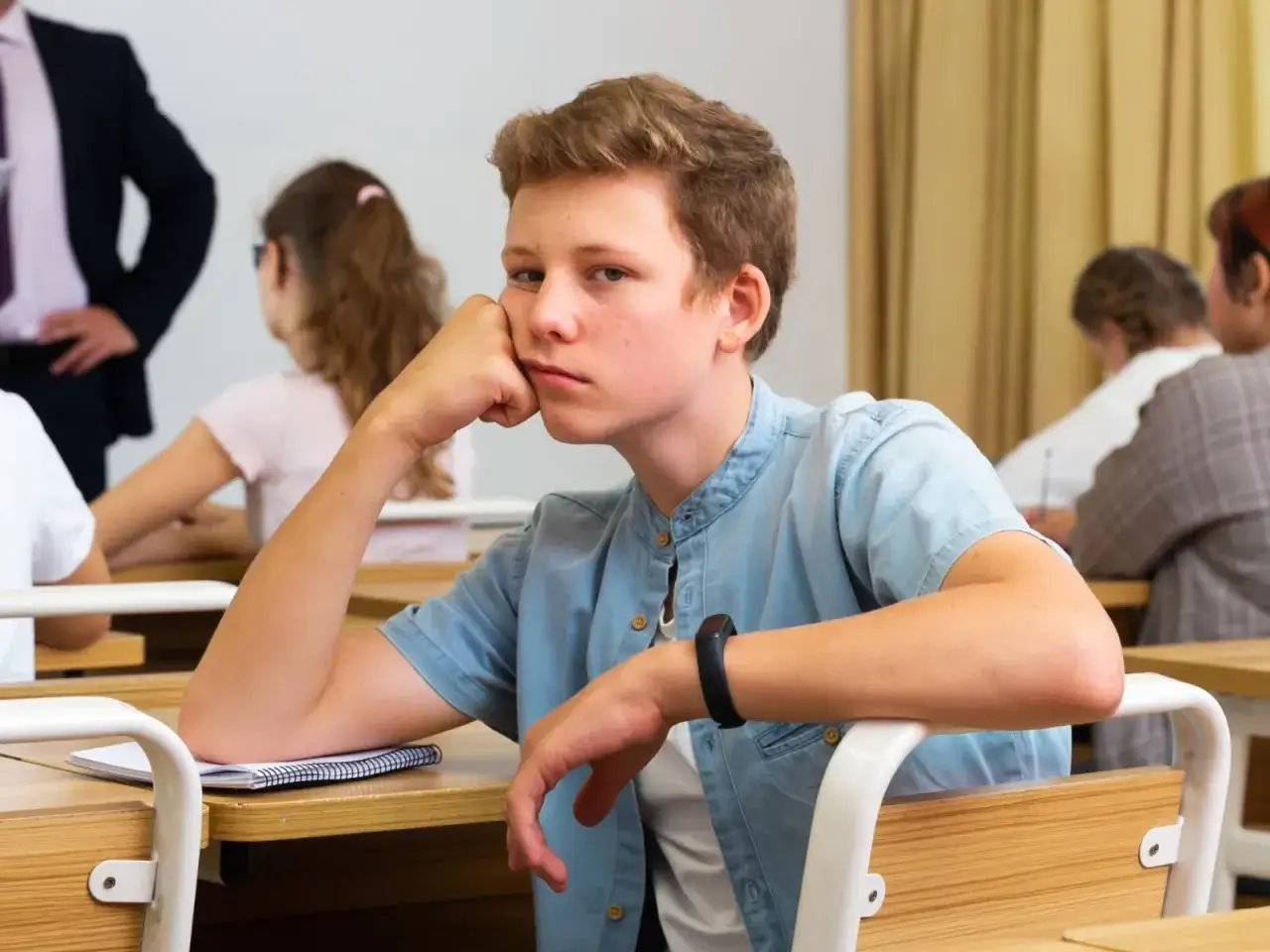 Znudzony uczeń opiera głowę na dłoni, podczas gdy wychowawca stoi z tyłu.