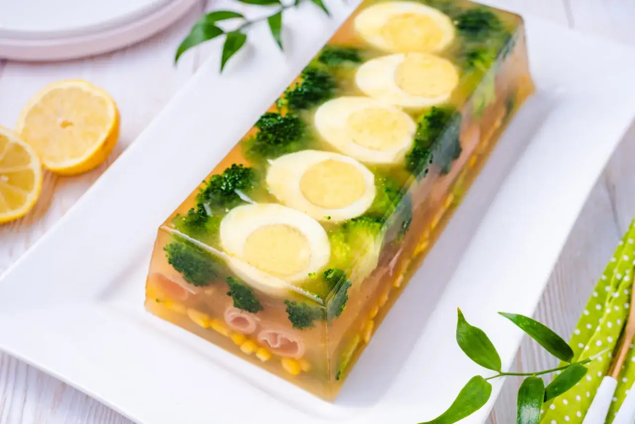 Eleganckie jajka w galarecie z brokułami i kukurydzą, ozdobione plasterkami cytryny.