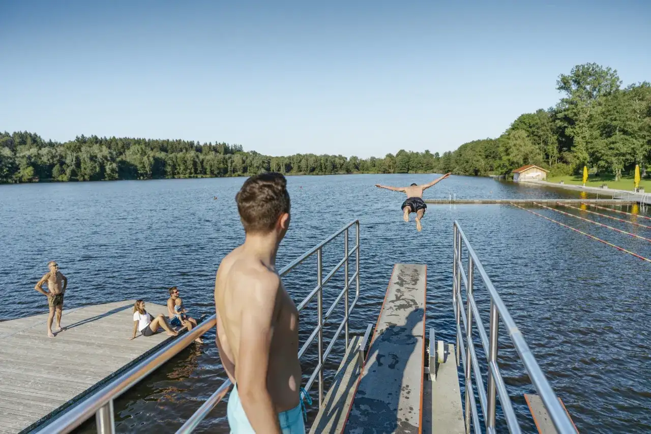 Ein Junge springt im Freibad Leutkirch von einem Sprungbrett ins Wasser. Andere genießen die Sonne am Ufer.