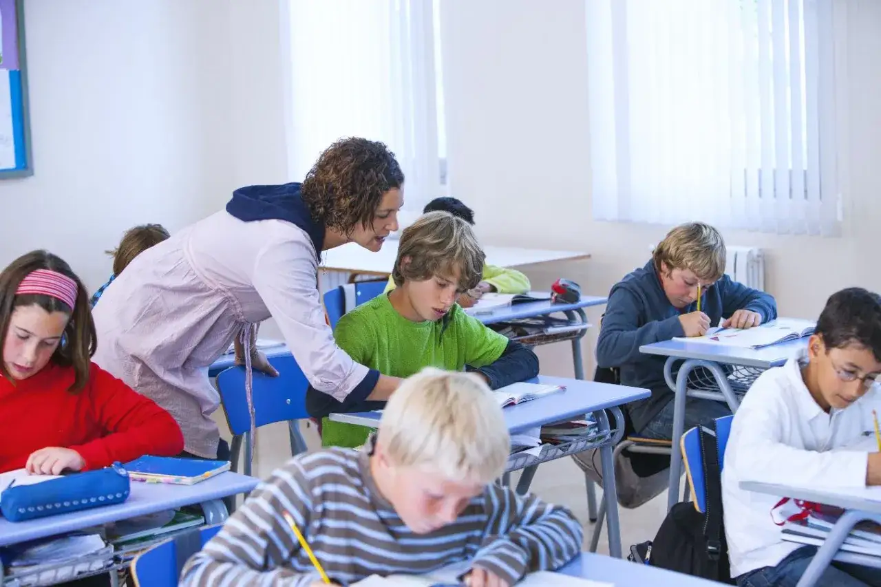 Nauczycielka pochyla się nad uczniem, pomagając mu w zadaniu. Dzieci piszą w zeszytach, zastanawiając się, ile wcześniej trzeba zapowiedzieć sprawdzian.