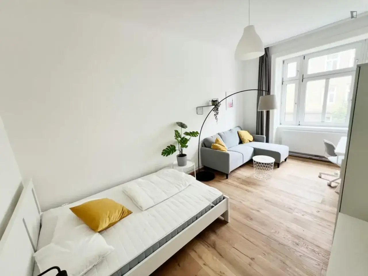 Chambre lumineuse pour logement étudiant à Vienne, Autriche. Lit simple, canapé gris, bureau et grande fenêtre.