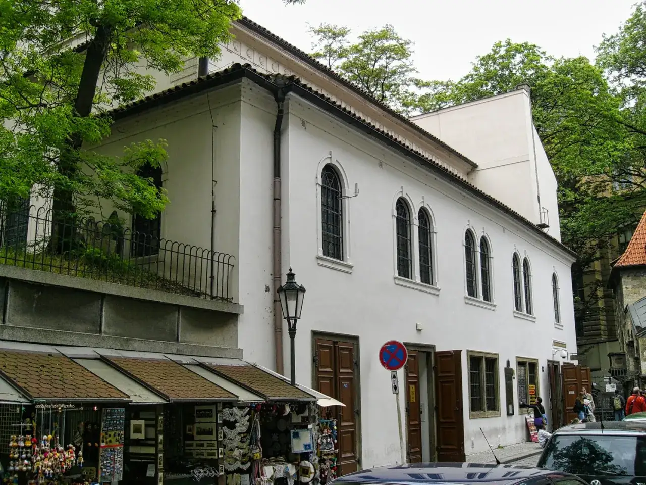 Die **Synagoge Prag** ist ein wei&szlig;es Geb&auml;ude mit Rundbogenfenstern. Davor ein kleiner Markt mit Souvenirs und ein Laternenpfahl.