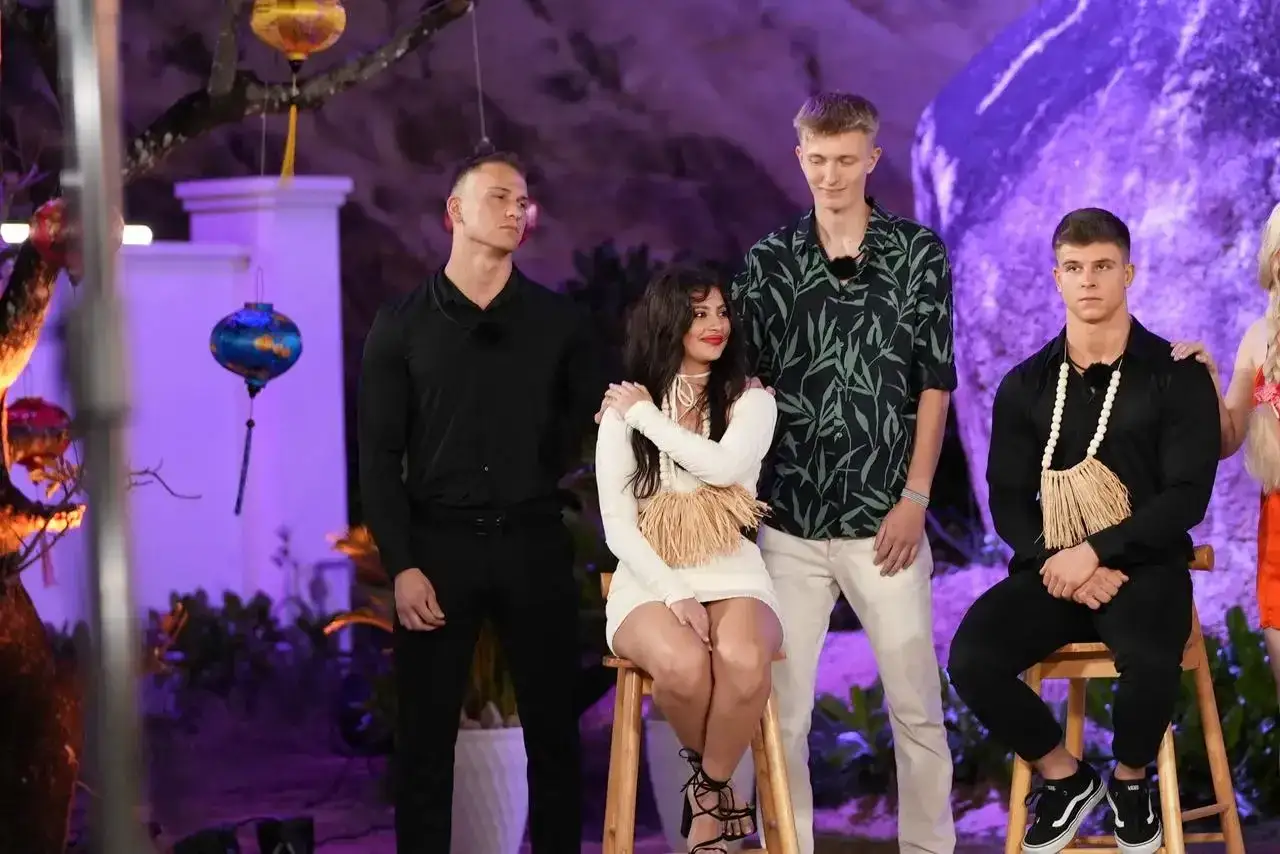 Hotel Paradise 8: Uczestnicy w tropikalnej scenerii. Młodzi ludzie, wśród nich kobieta w białej sukience, czekają na rozwój wydarzeń.