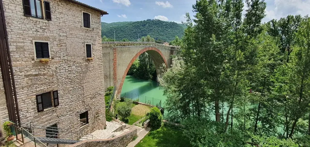 Un antico ponte ad arco domina un fiume verde smeraldo, perfetto per il trekking nelle Marche. Un borgo in pietra completa la scena.
