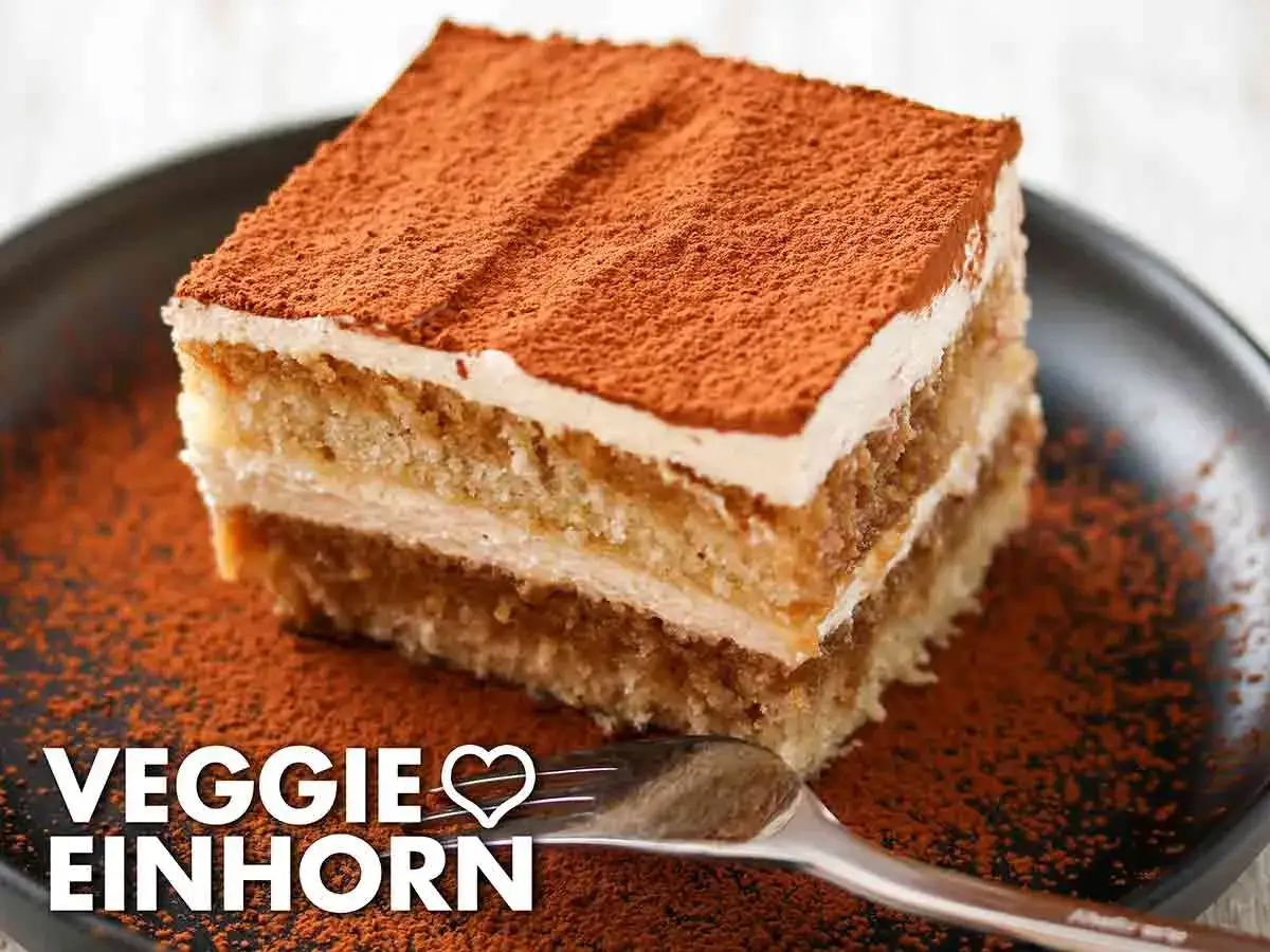Ein Stück veganes Tiramisu ohne Alkohol, bestäubt mit Kakao, auf einem Teller mit Kakaopulver und einer Gabel.
