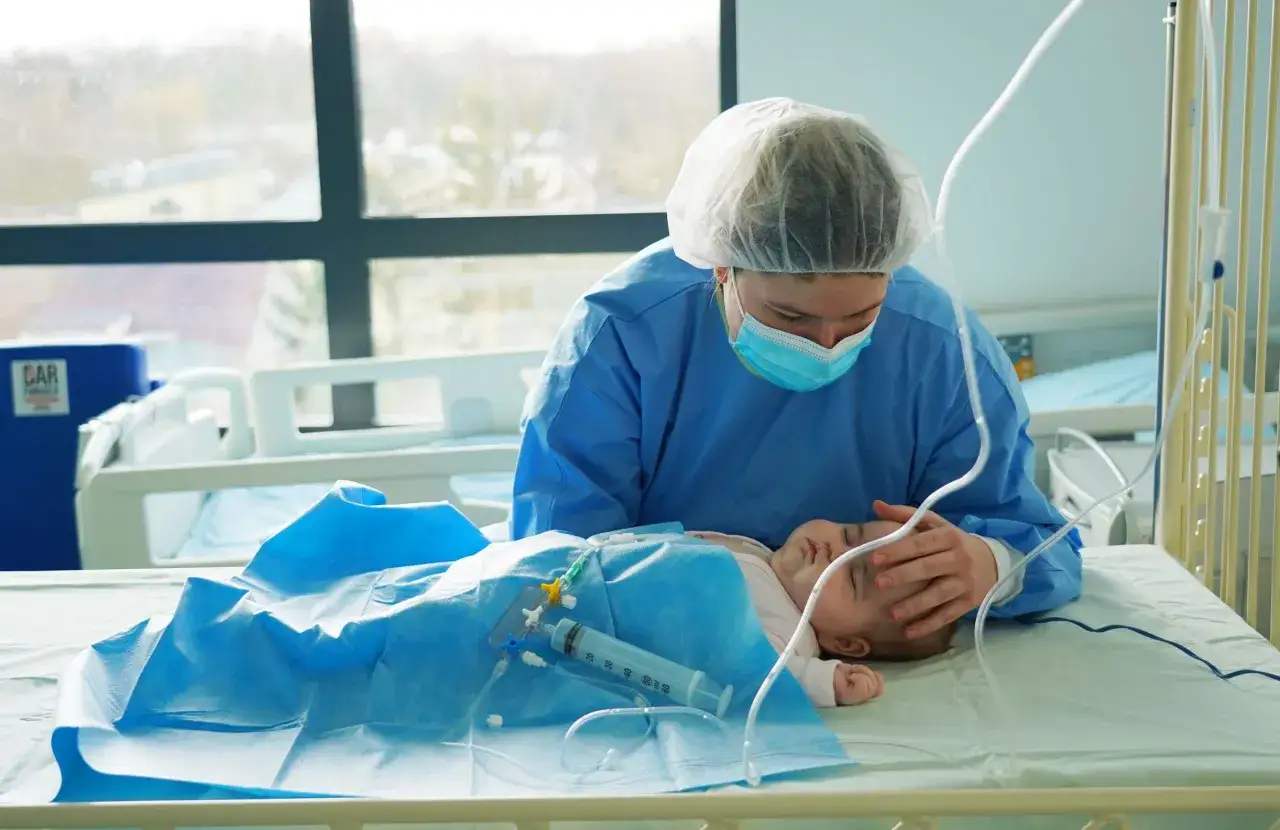 Opiekunka w niebieskim fartuchu i masce pochyla się nad śpiącym dzieckiem z si. W tle szpitalne łóżka i okno.