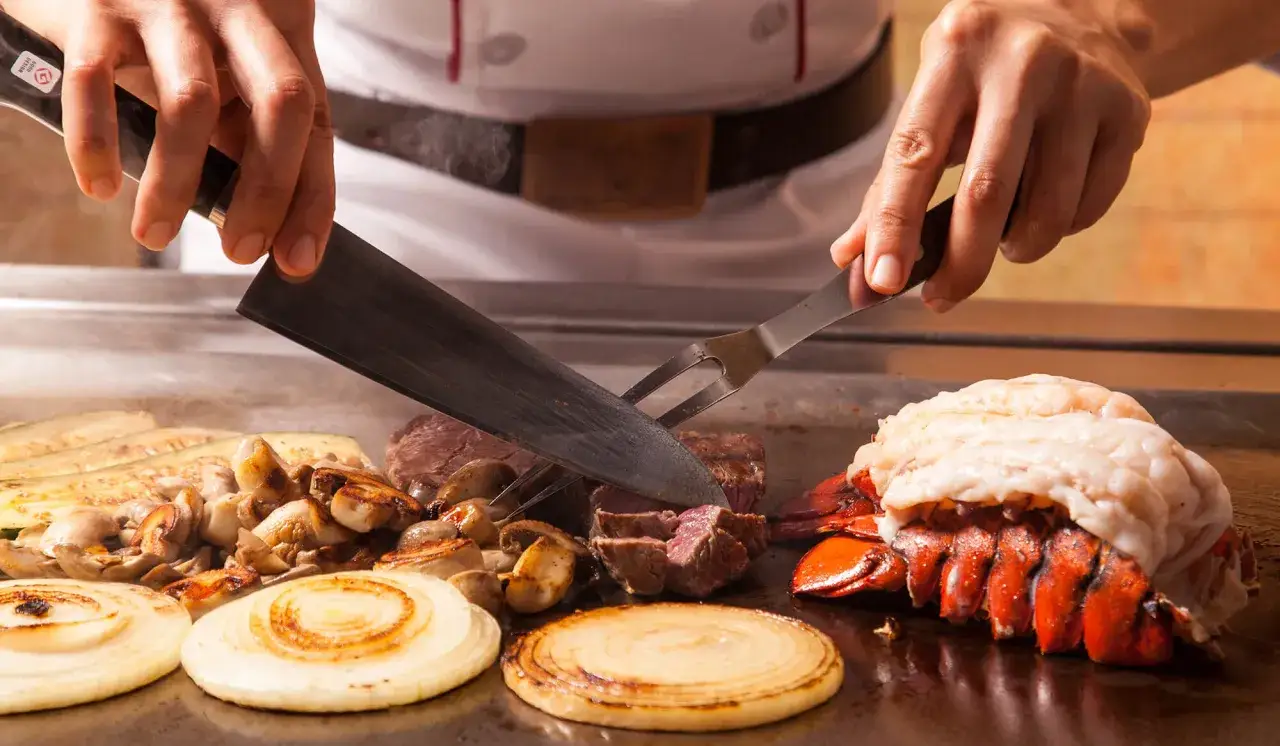 Chef corta carne y langosta en una plancha caliente, mostrando la preparación de teppanyaki que es. Verduras y champiñones se cocinan al lado.