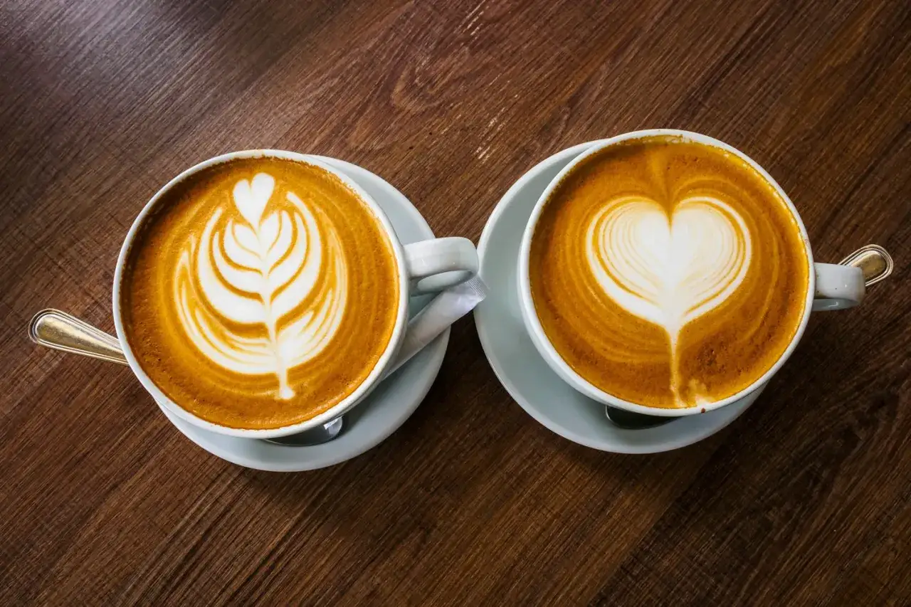 Tv&aring; kaffekoppar med latte art p&aring; tr&auml;bord. En med ett l&ouml;vm&ouml;nster, den andra med ett hj&auml;rta. En visuell skillnad mellan latte och cappuccino.
