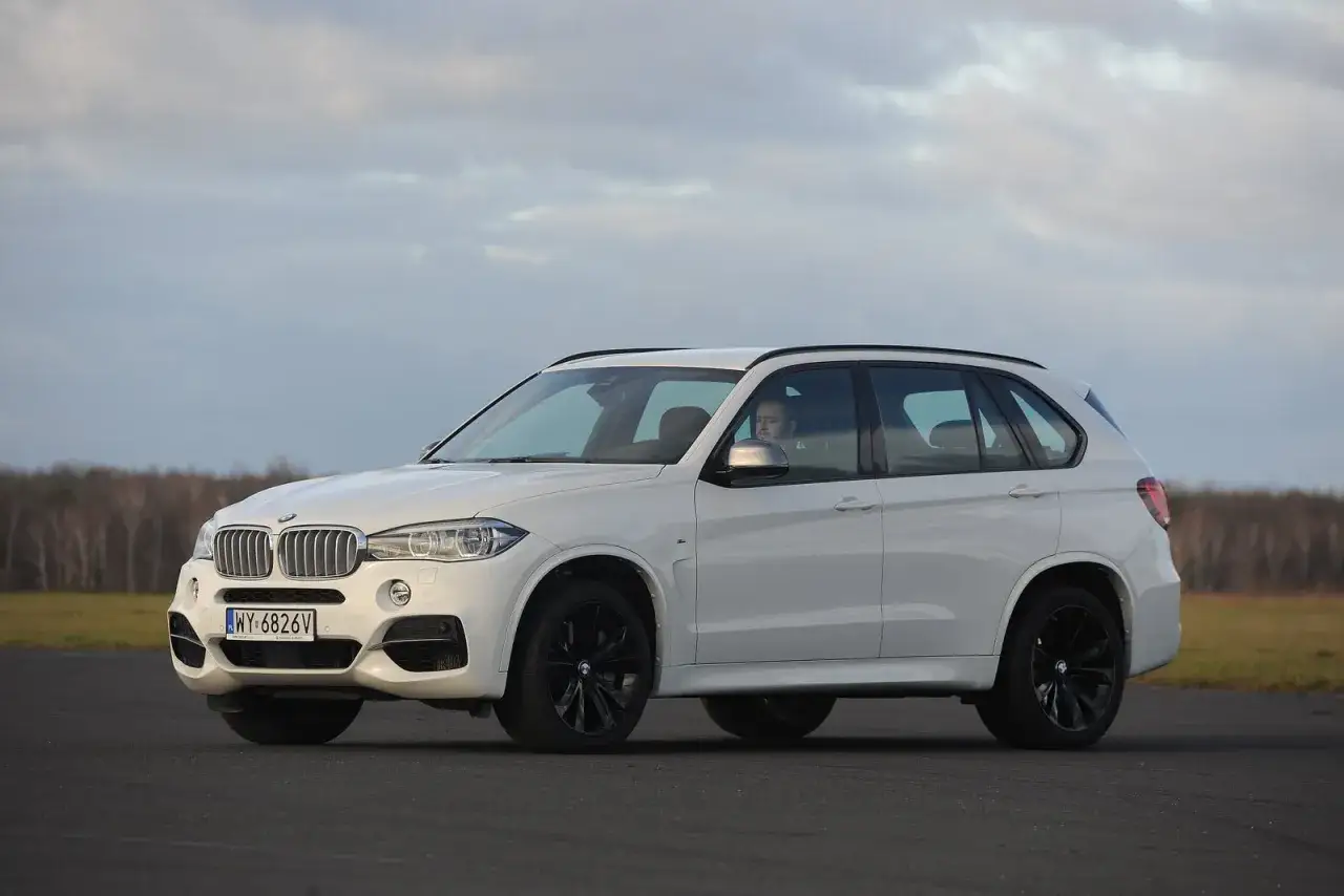 Białe BMW X5 M50d z czarnymi felgami na tle pochmurnego nieba i drzew.