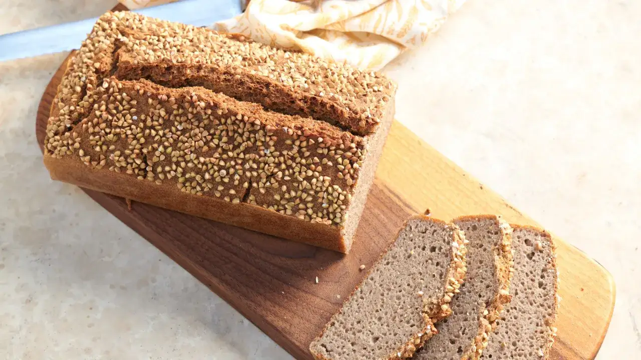 Ein Laib **Buchweizen**brot, bestreut mit Buchweizenk&ouml;rnern, liegt auf einem Holzbrett. Daneben liegen drei Scheiben Brot.