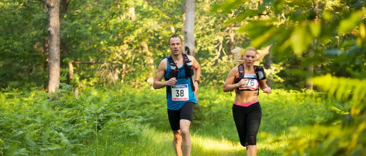 Zwei L&auml;ufer, Mann und Frau, nehmen an einem **Ultramarathon Deutschland** teil. Sie rennen durch einen sonnendurchfluteten Wald mit Farnen und B&auml;umen.