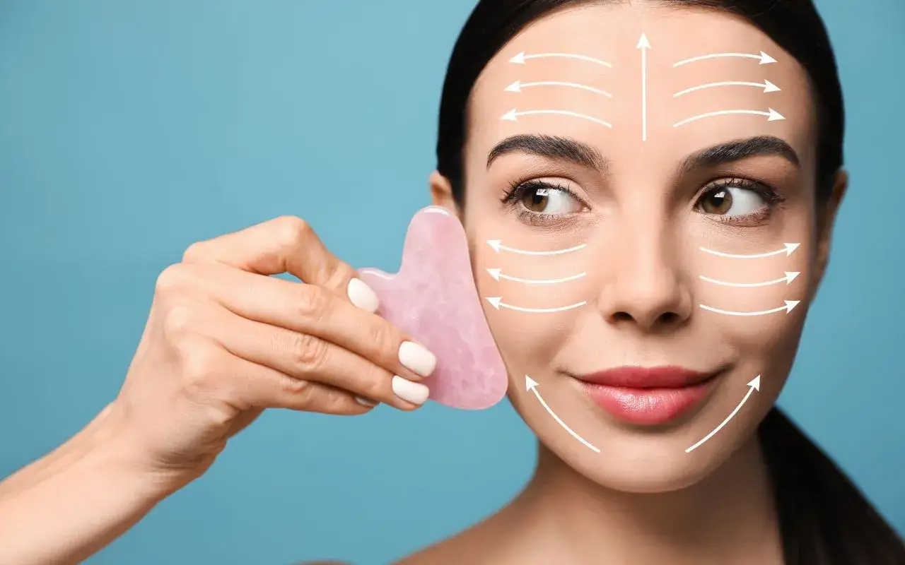 Donna usa un gua sha viso rosa per un massaggio tonificante, con frecce bianche che indicano i movimenti.
