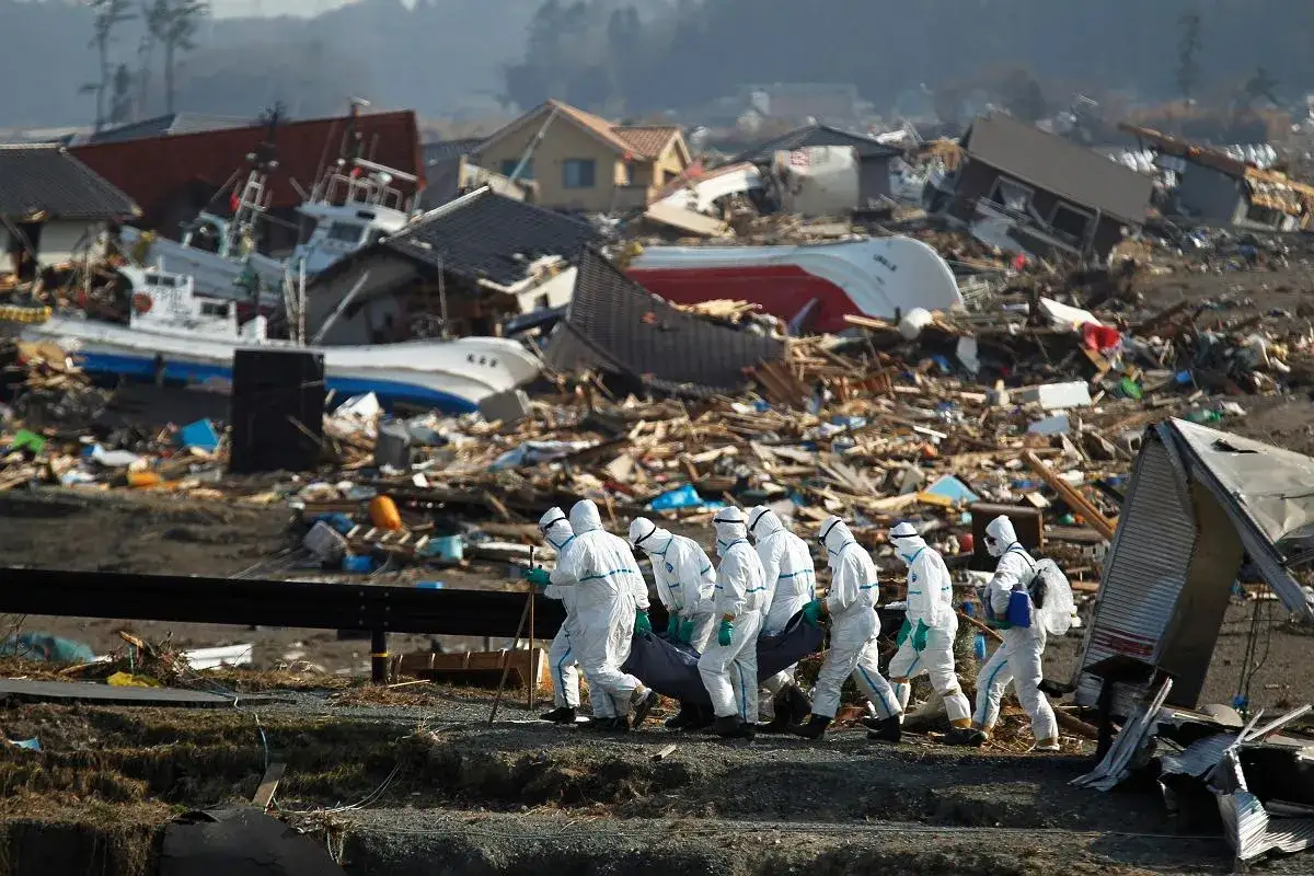 Ratownicy w kombinezonach ochronnych niosą ciało przez zniszczone miasto po katastrofie w Fukushimie.