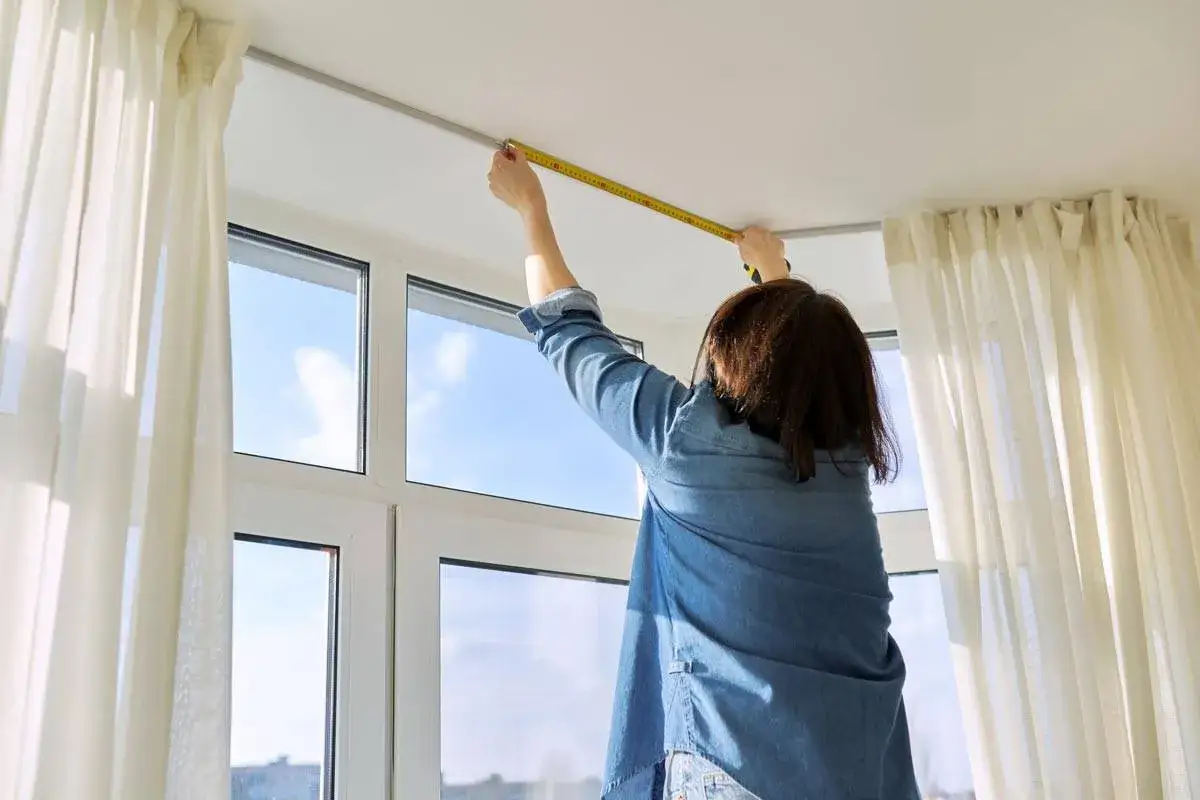 Persona misura l'altezza del bastone per tende dal soffitto, vicino a una finestra con tende bianche.