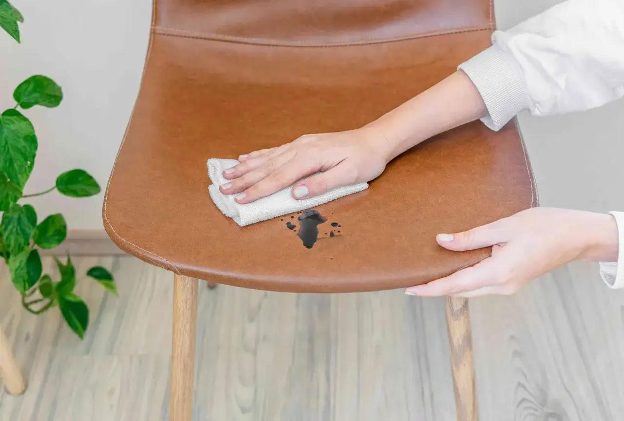 Mano che pulisce una macchia scura su una sedia in pelle marrone con un panno bianco, per smacchiare pelle.