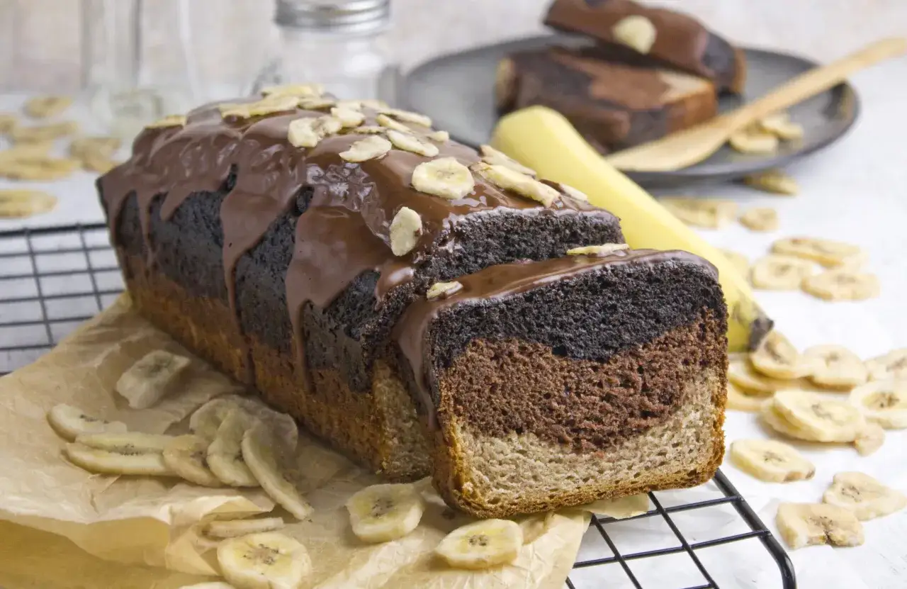 Ein saftiges Bananenbrot mit Schokolade, garniert mit Bananenchips, liegt auf einem Gitter. Daneben liegen getrocknete Bananenscheiben.