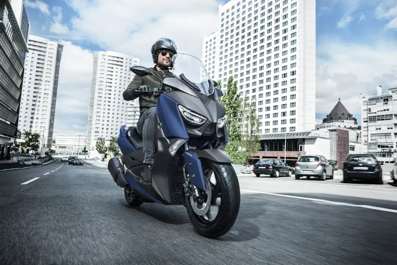 Mężczyzna w kasku i okularach przeciwsłonecznych jedzie niebieskim skuterem Yamaha X-MAX 400 po miejskiej drodze. W tle wysokie budynki i samochody.